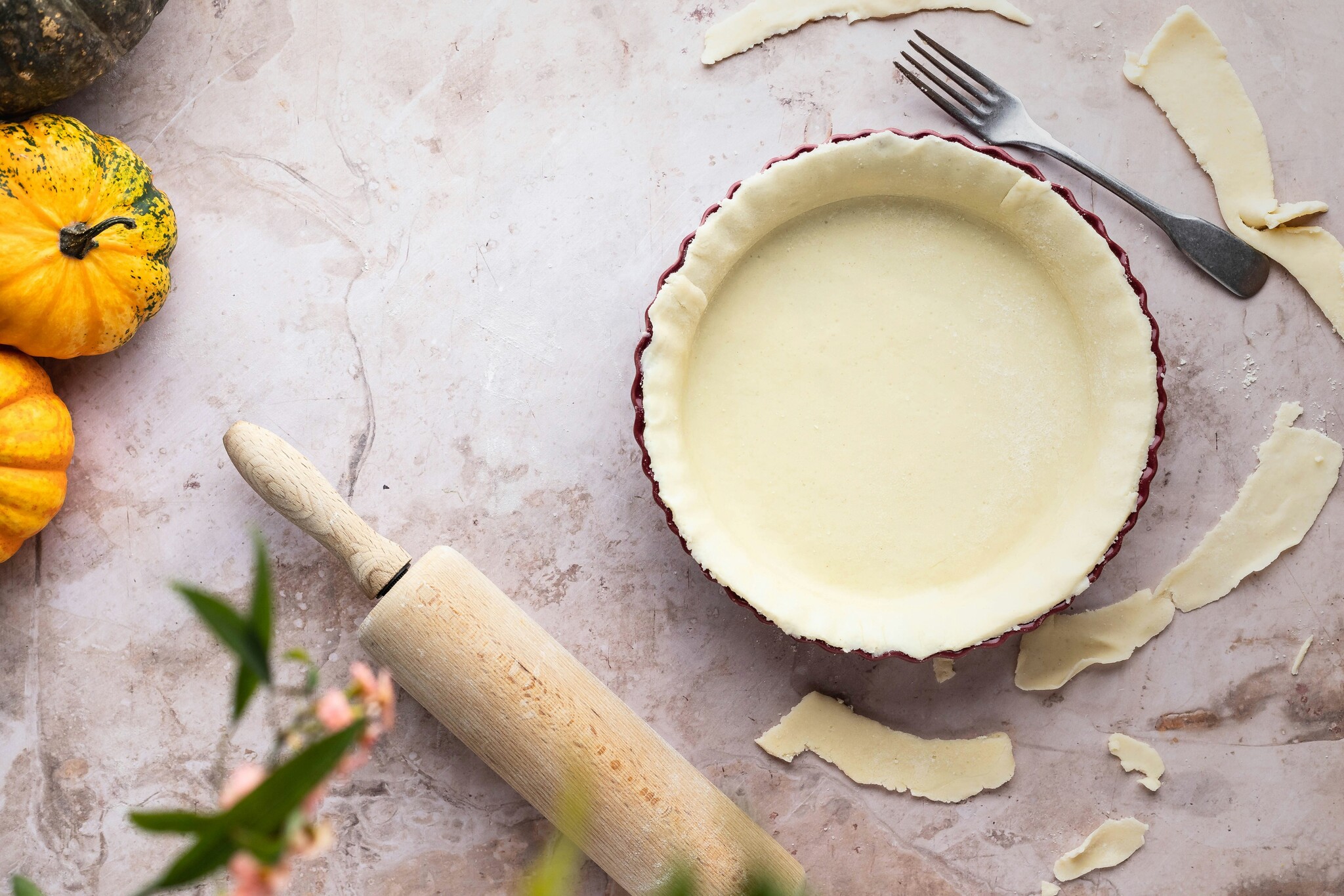 a pie crust sits inside a pie pan with excess dough, a fork, a rolling pin, and small pumpkins spread around it. 