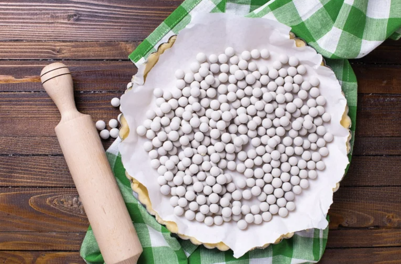 white, circular pie weights sit inside a parchment-lined pie crust on a wooden tabletop with a rolling pin and green plaid towel beside it. 