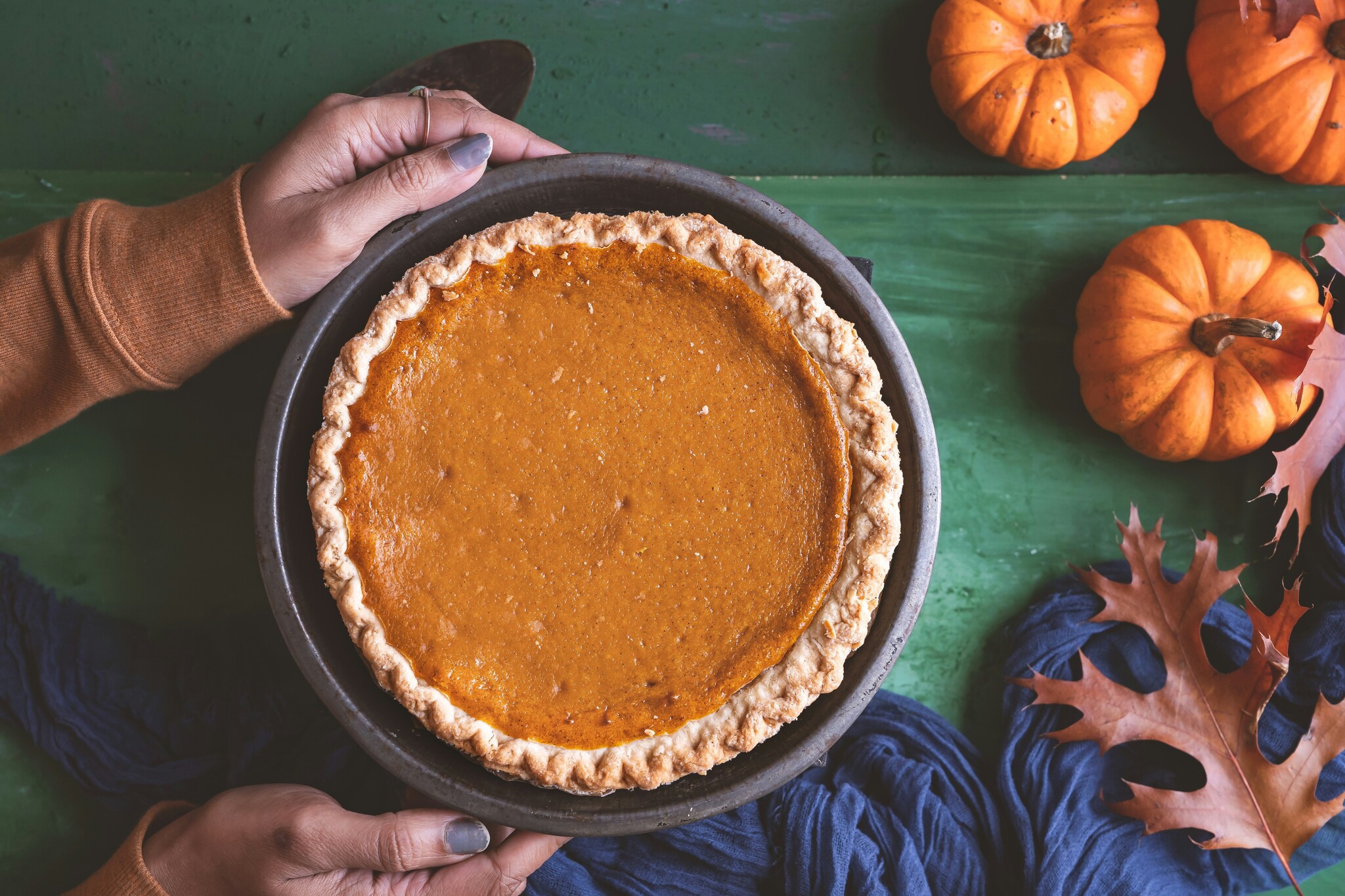 A person holds a pumpkin pie over a green surface with mini pumpkins and fall leaves surrounding it. 