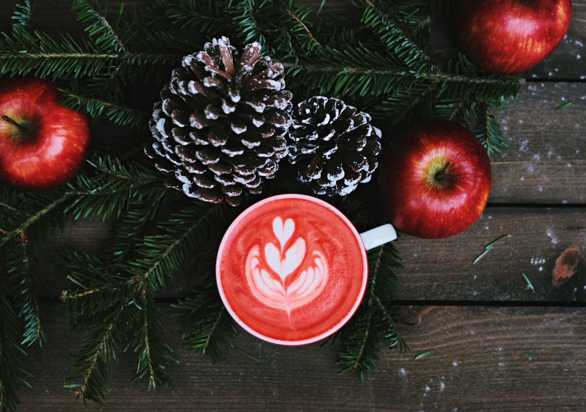 A red latte sits on a wooden table with snow-coated pine cones, red apples, and pine branches around it. 
