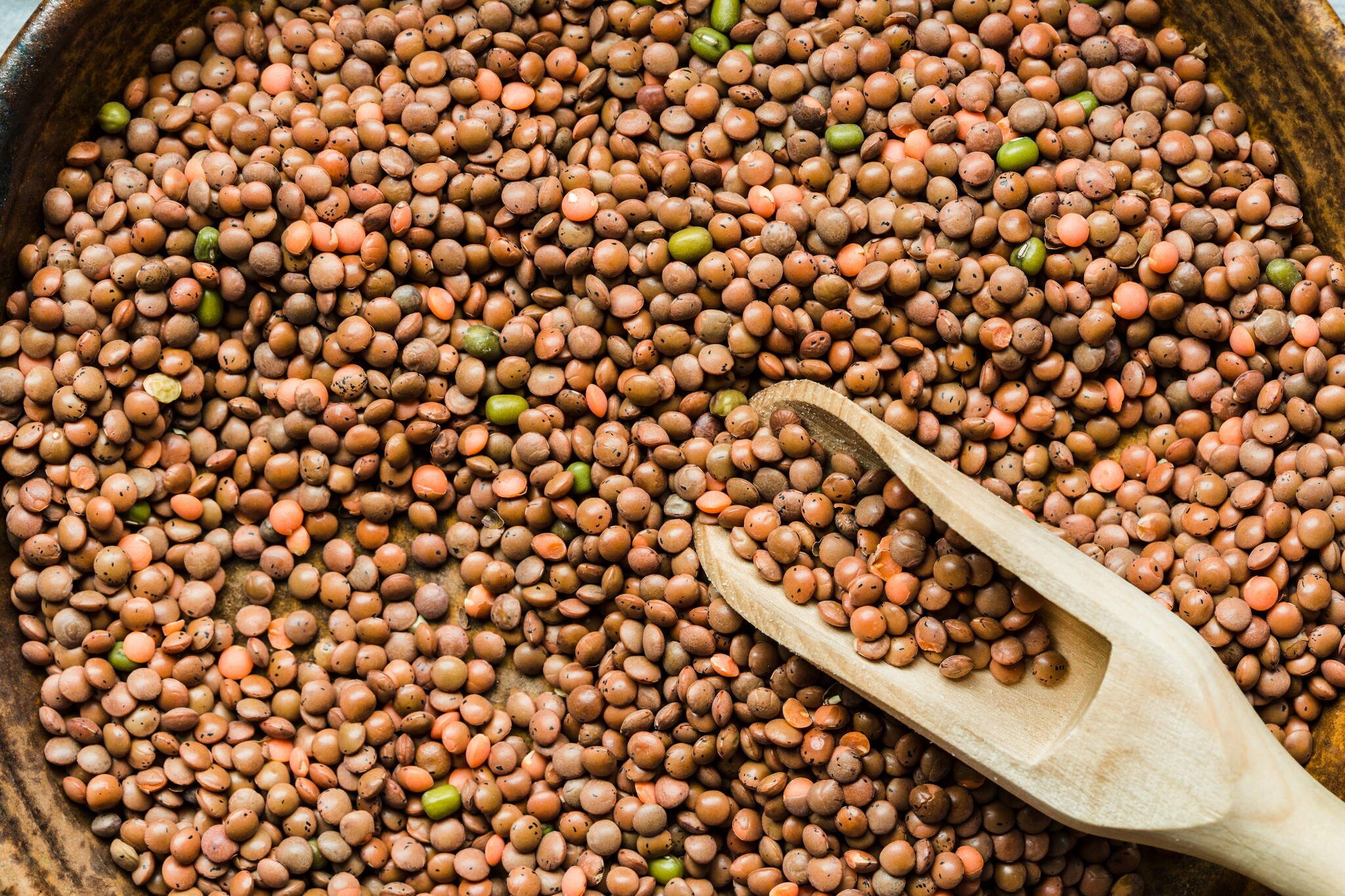 Reddish-brown lentils sit in a large bowl with a wooden scoop. 