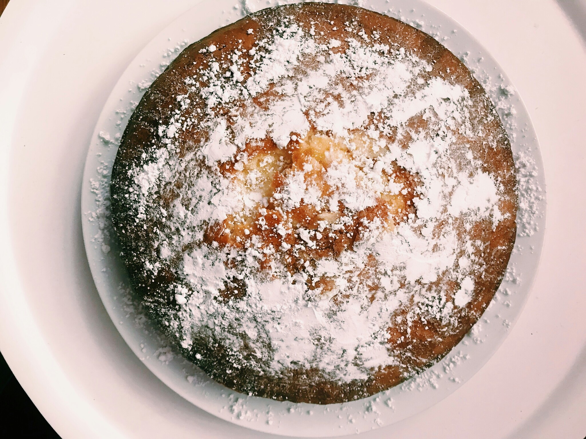 A round cake covered in powdered sugar sits on a white plate on a white countertop. 