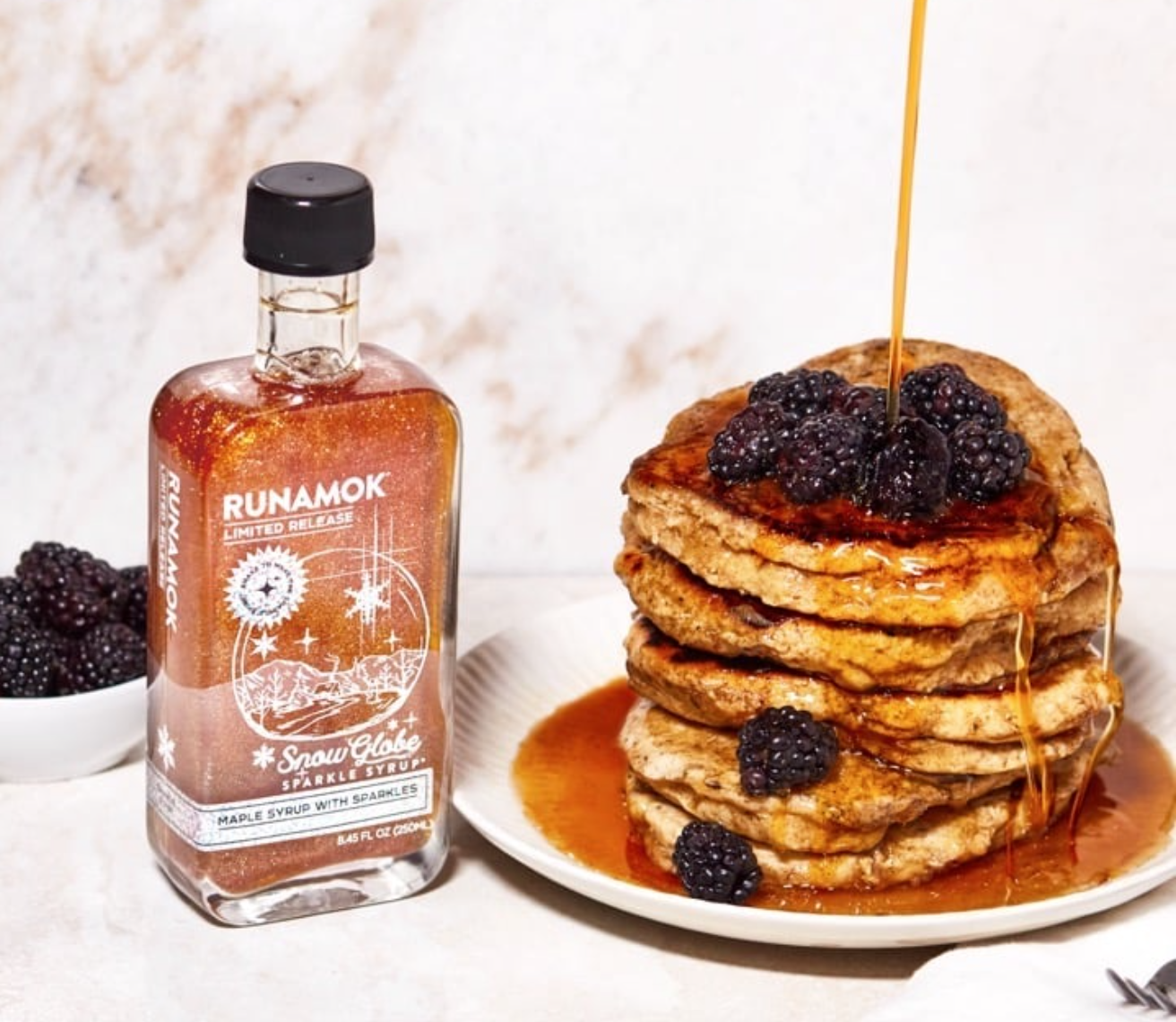 A bottle of Runamok Sparkle Syrup sits beside a stack of syrup-drenched pancakes topped with blackberries. 