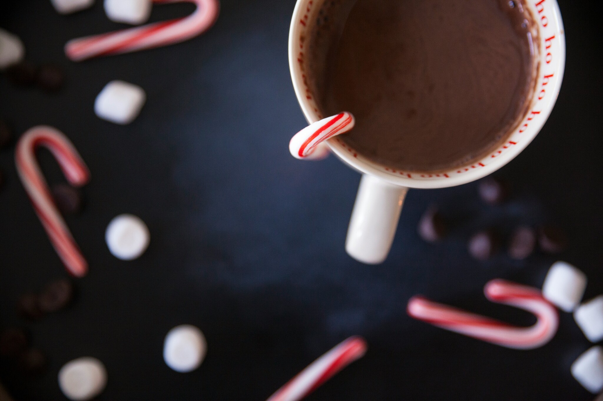 A cup of coffee with a candy cane hanging from it sits on a black surface scattered with candy canes and marshmallows.