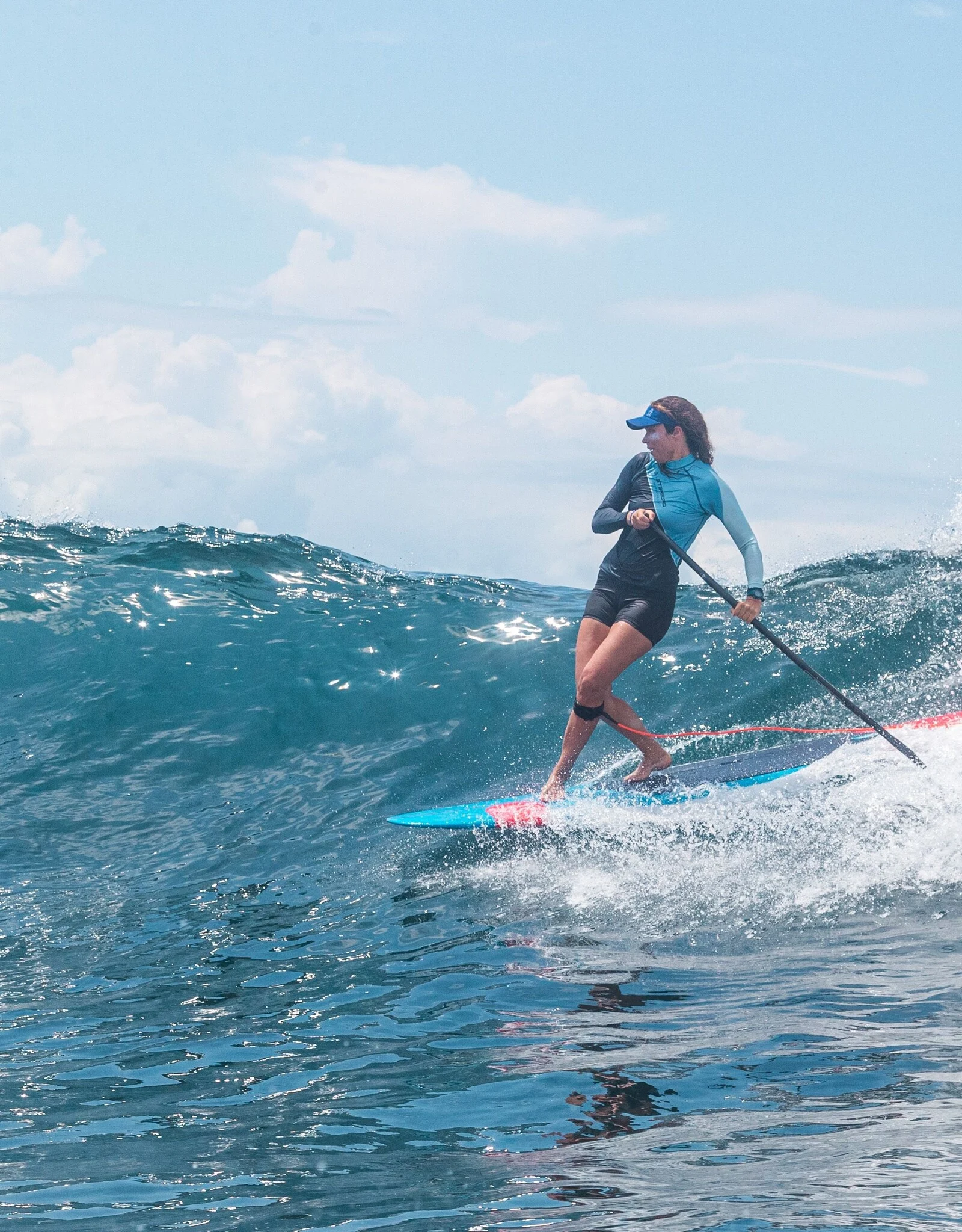 Woman carving down the face of a small wave on a blue longboard SUP, paddle in hand, with a visor and long-sleeve top under a bright sky.