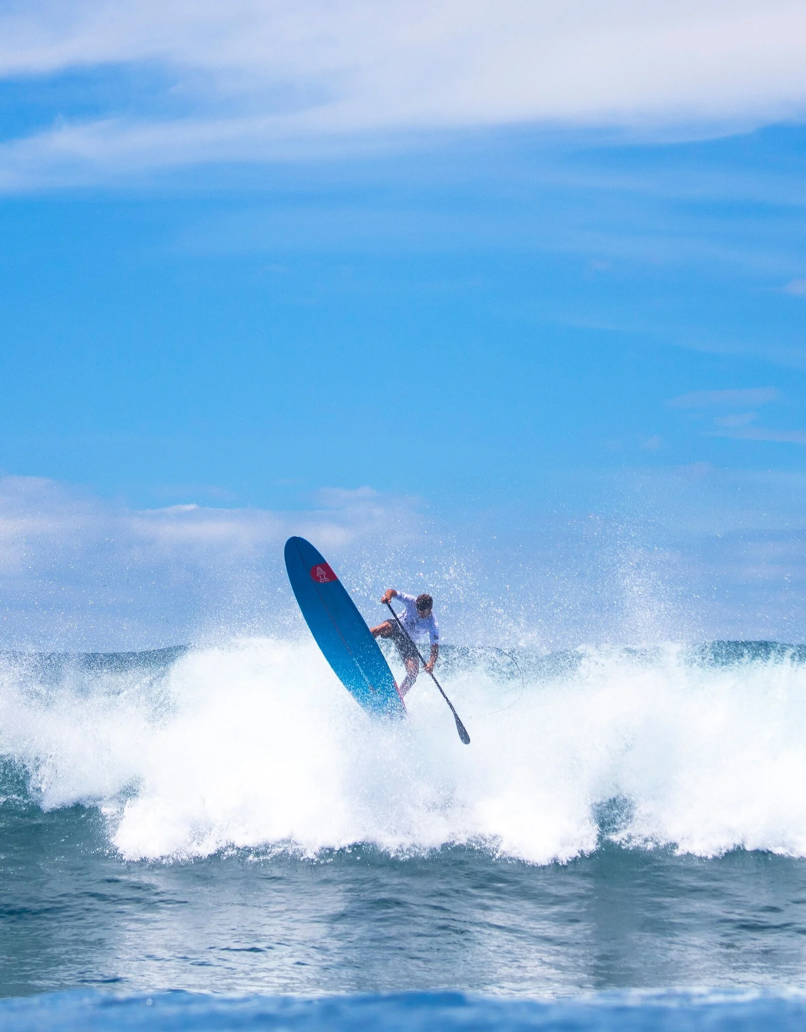 SUP surfer launching off the lip of a powerful wave on a blue board, caught mid-air against a bright blue sky with white spray below.
