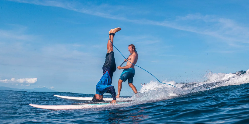 Two riders having fun on long Starboard Go Surf boards, one towing the other while doing a headstand.