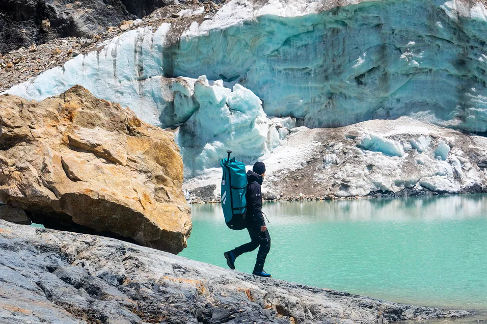 Hiker carrying a Fanatic Fly Air Pocket Edition inflatable SUP backpack along a turquoise glacial lake.
