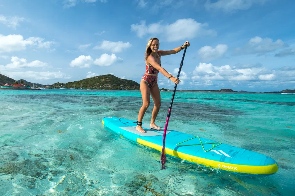 Woman standing and paddling a Fanatic Fly Air Pocket Edition inflatable SUP in shallow, glassy turquoise water.