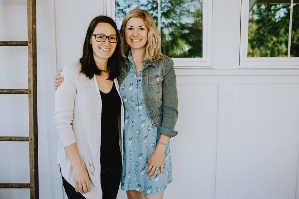 Friendly Patricia and Ashley, owners of Hansell and Halkett are standing beside each other and smiling. A minimalistic white wall is in the background with a bright window and rustic ladder off to the side..