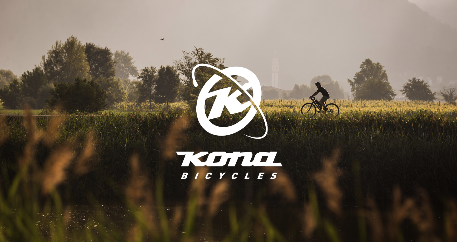 Kona Bicycles logo superimposed over a person cycling on their Kona bike through a grassy field on a sunny day.