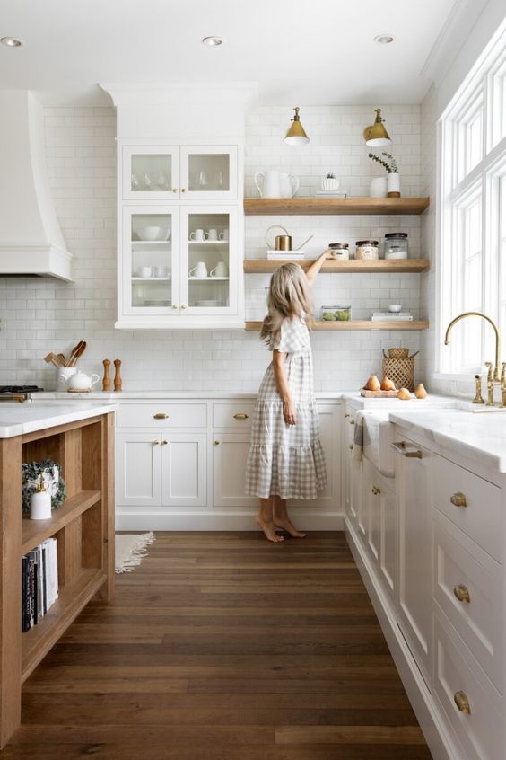 Wood shelves sit above white cabinets in a bright open kitchen. A woman is reaching for an item off one of the shelves.