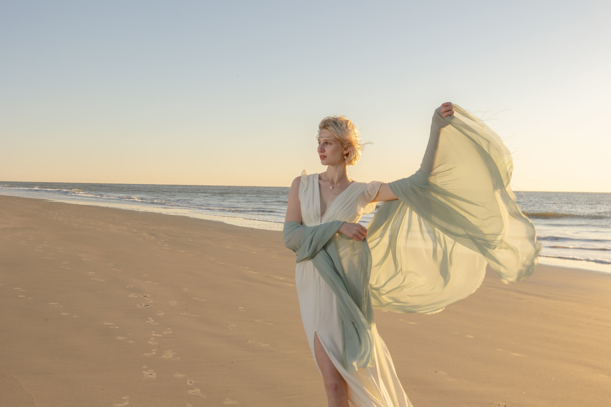 A white woman with short, white-blonde hair stands on a beach at golden hour, tossing her seafoam green shawl into the air to catch the wind. 