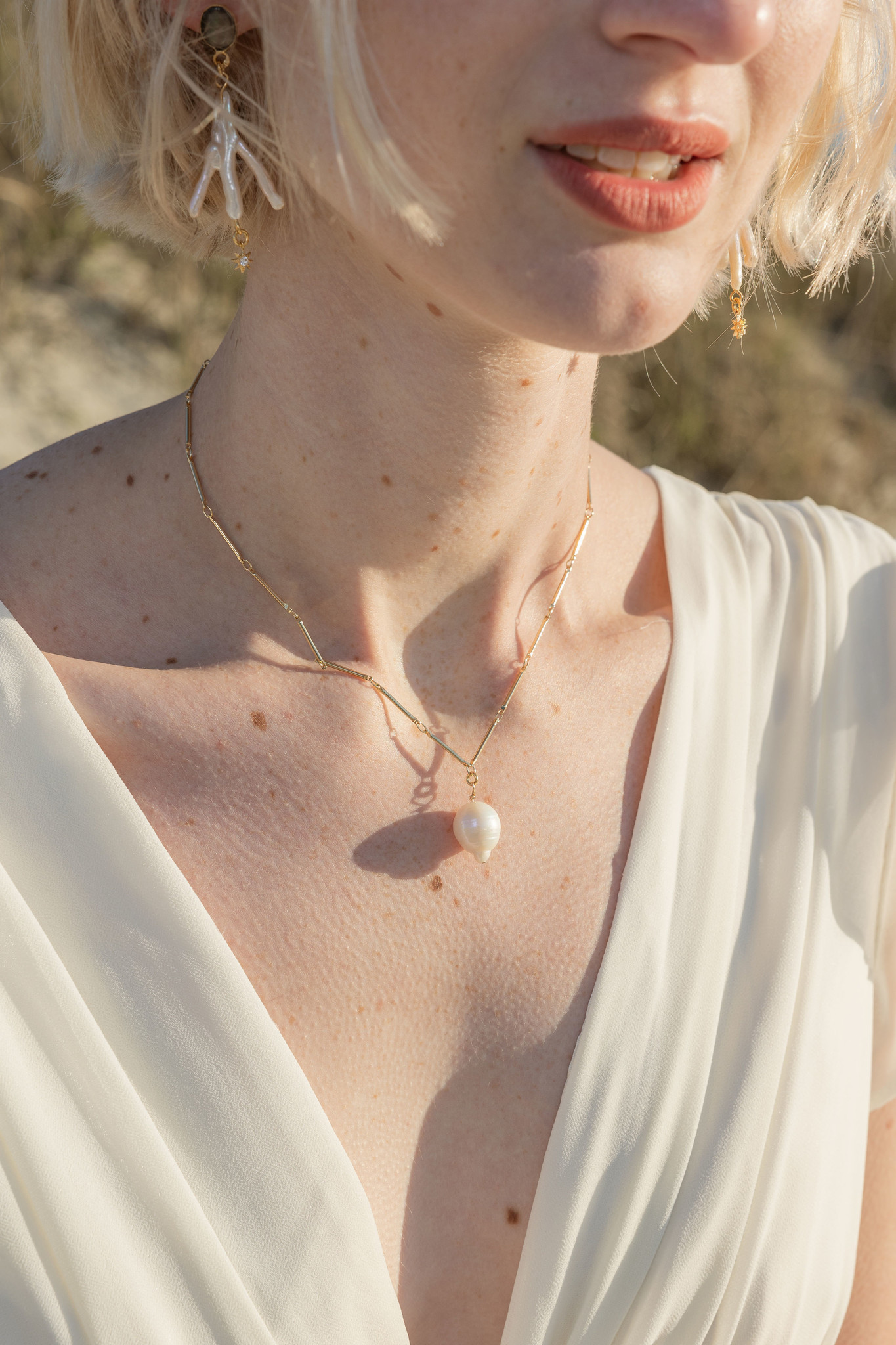 A white woman with white-blonde hair and myriad freckles smiles softly in the late-afternoon sunlight. A gleaming freshwater pearl earring dangles from her ear, branched like a chicken foot with a dark stone at the top and a tiny, golden star hanging at the bottom. Around her neck is a gold, stick-chain necklace holding a single, freshwater pearl. 