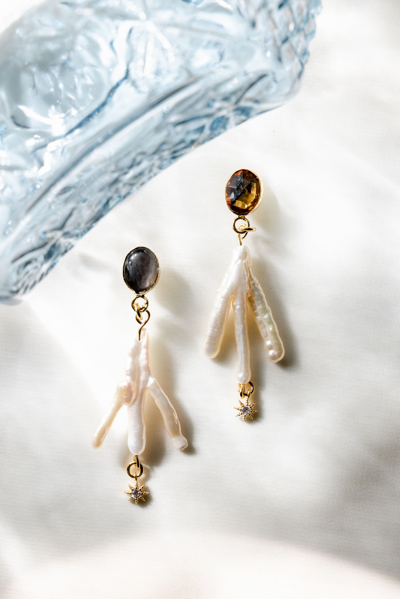 A pair of gleaming, freshwater pearl earrings shaped like chicken feet or coral with three branches. The bottoms hold tiny golden stars, while the top is secured by a topaz on one, and another stone on the other. 