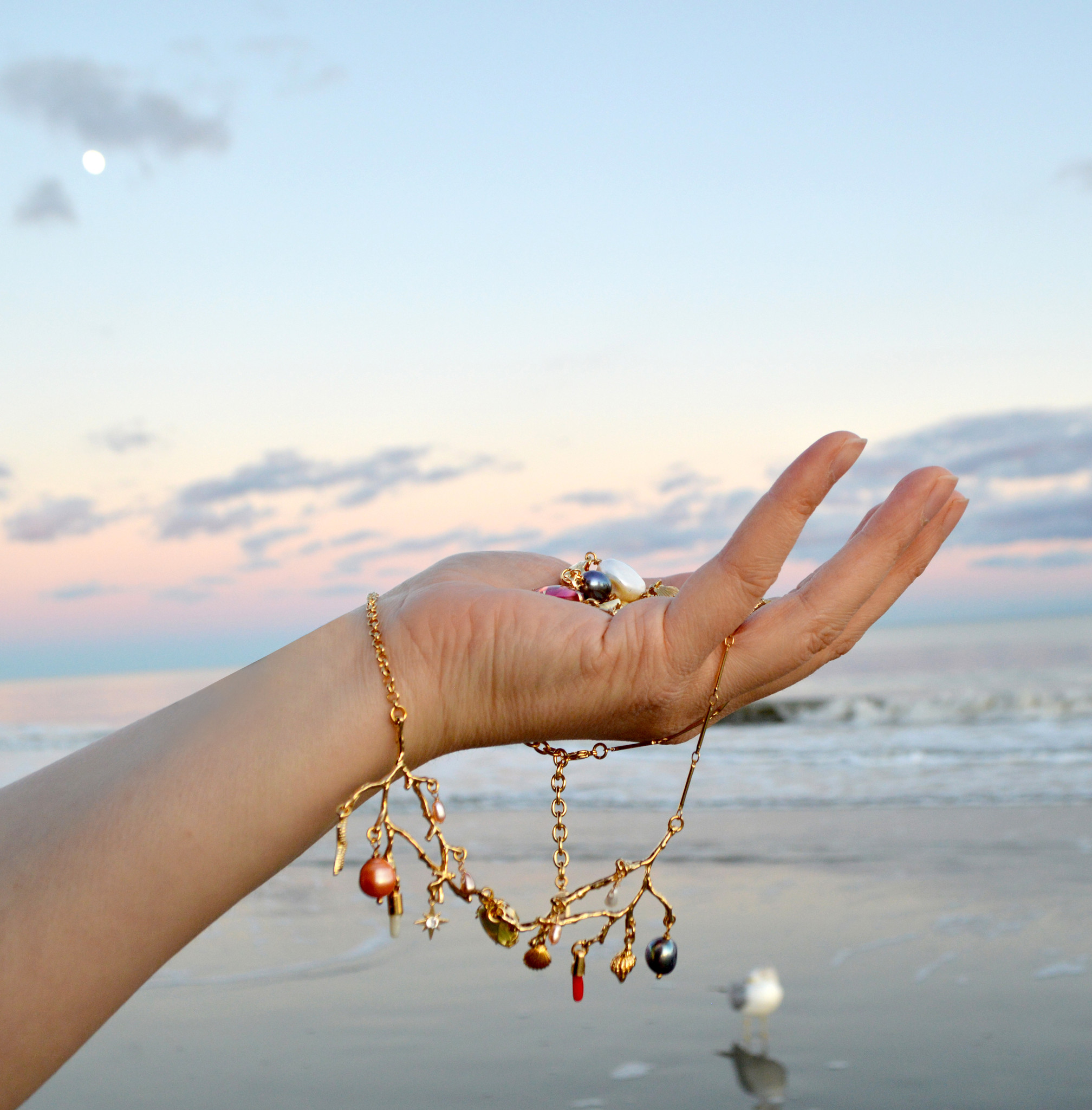 A light-skinned hand holds multiple pieces of jewelry including a necklace that dangles from the bottom, shaped like golden coral branches covered in seashells, coral-shaped charms, freshwater pearls, etc. It's set against the backdrop of an East coast beach at dusk, with soft pink, blue, and purple skies and a calm sea, the full moon in the top left corner. 