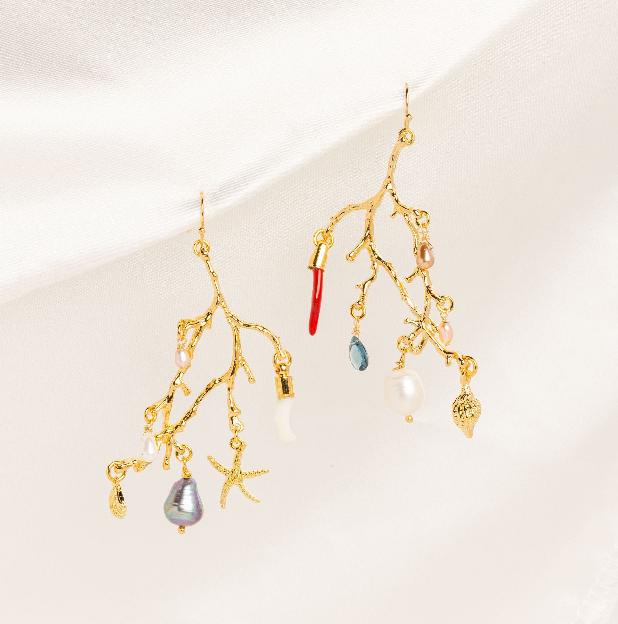A pair of gleaming coral-branch earrings from which dangle tiny gold seashell charms, glass stones, coral-shaped charms, and freshwater pearls. They are set against a cream-colored background.