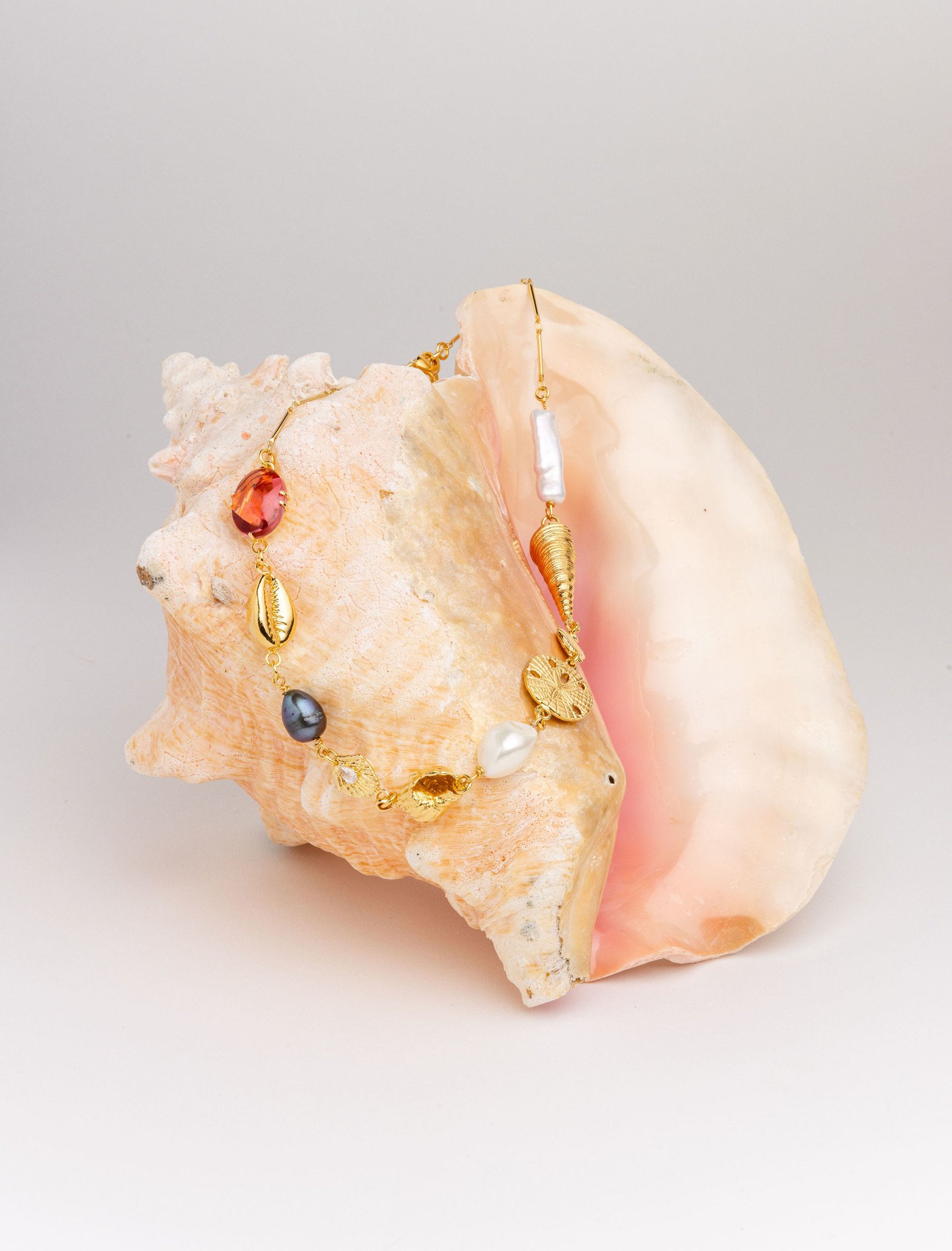A delicate charm necklace with golden seashells, sand dollars, pearls, and glass stone jewels is draped over a brilliant conch shell with a soft pink interior and peachy-gold exterior. 