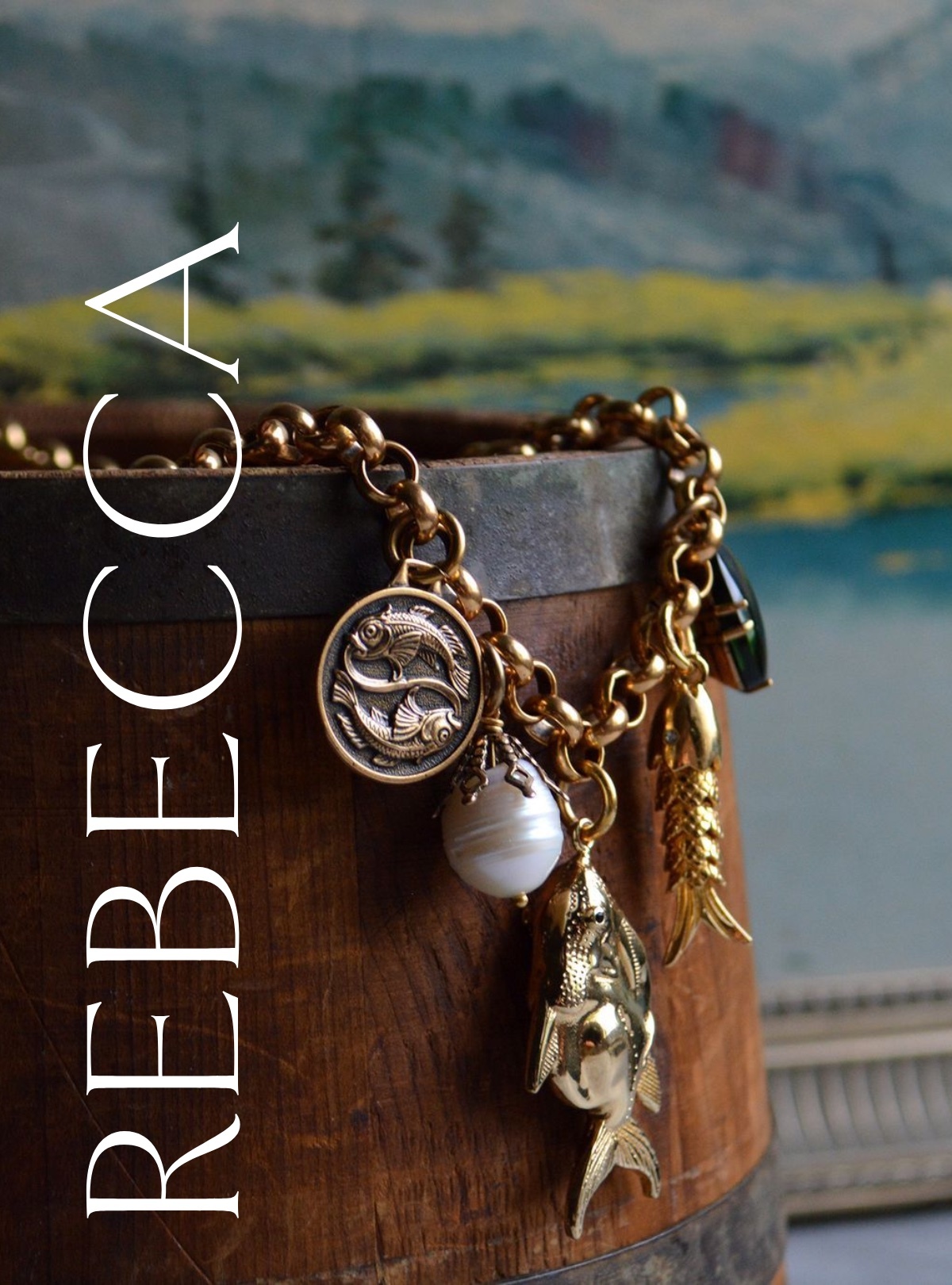 Bright gold charms hang on a thick, rolo chain: two fish circling one another in the shape of Pisces, a pearl, an articulated fish, and a green glass stone. They're draped on an antique bucket in front of a soft blue-green painting that serves as the backdrop. REBECCA is written down the left side in white.