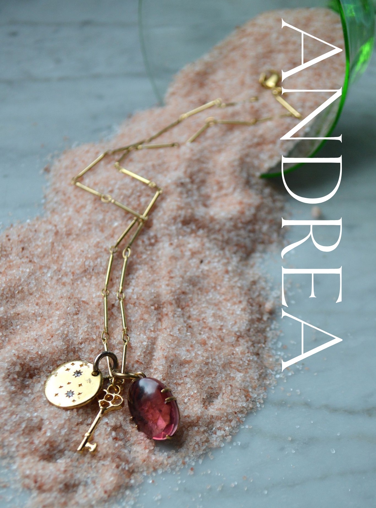 A light green glass spills pink himalayan salt across a marble counter. In it lies a golden stick chain with a bright gold key charm, a small circular one with tiny crystals set like stars, and a rosy-pink glass stone set in antiqued gold.