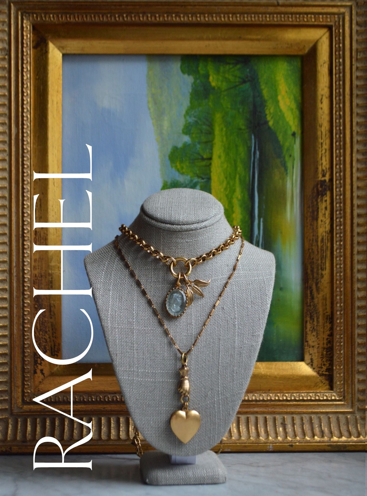 A grey, headless bust holds two gleaming gold chains with a small, oval blue cameo charm and a charm made of three pointed leaves. The bottom necklace is made of a heart-locket held by a knight's gauntlet. It's set against a gold-framed painting of a blue, green, and white landscape. RACHEL is written on the bottom left side.