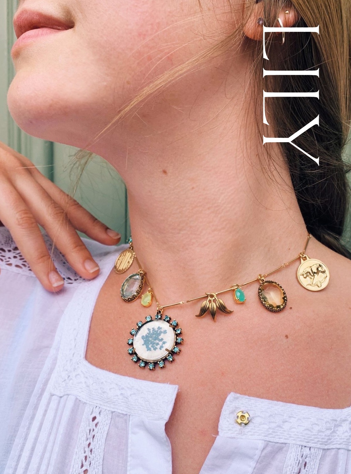 A white woman stands with her face turned to the side, lips, jaw, and hand in view of the camera. A stick chain necklace hangs from her throat, covered in eclectic charms from a central spray of blue flowers trapped in glass and surrounded by crystals, to other small, brightly-colored stones between gold ones.