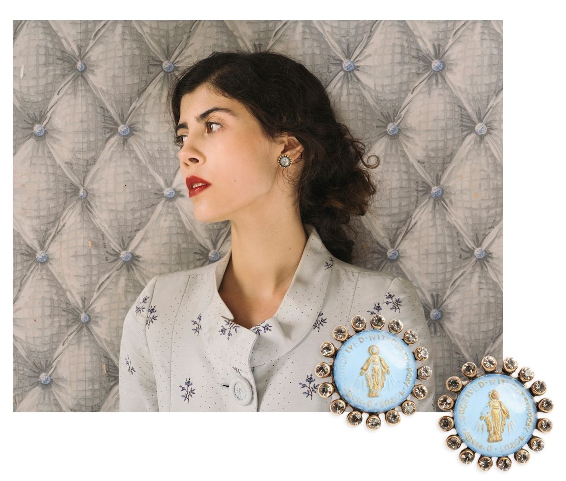 A white woman with dark hair in a low bun looks to the side, tears in her eyes. She's wearing a pair of light blue, circular earrings framed by tiny crystals, bearing a central motif of a religious figure with hands spread low in a holy stance. The earrings are seen closer up to the bottom right of her.