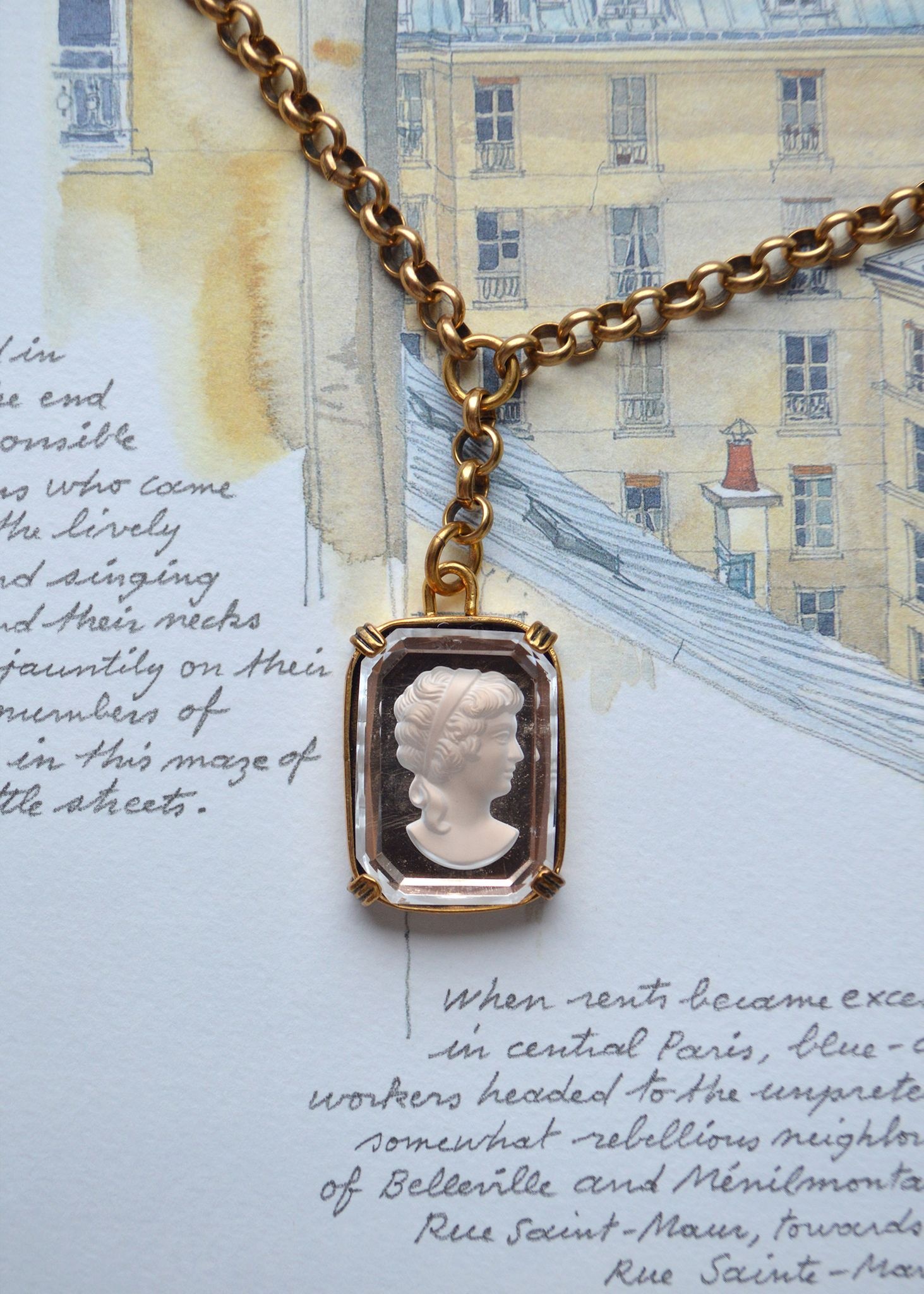 A chain of large, antiqued gold circles lies over a watercolor illustration of Paris, and delicate hand-written script. At the center of the necklace chain is a rectangular bobble covered in glass, depicting the cameo profile of a woman with loose, curly hair looking to the side against a darker background. 