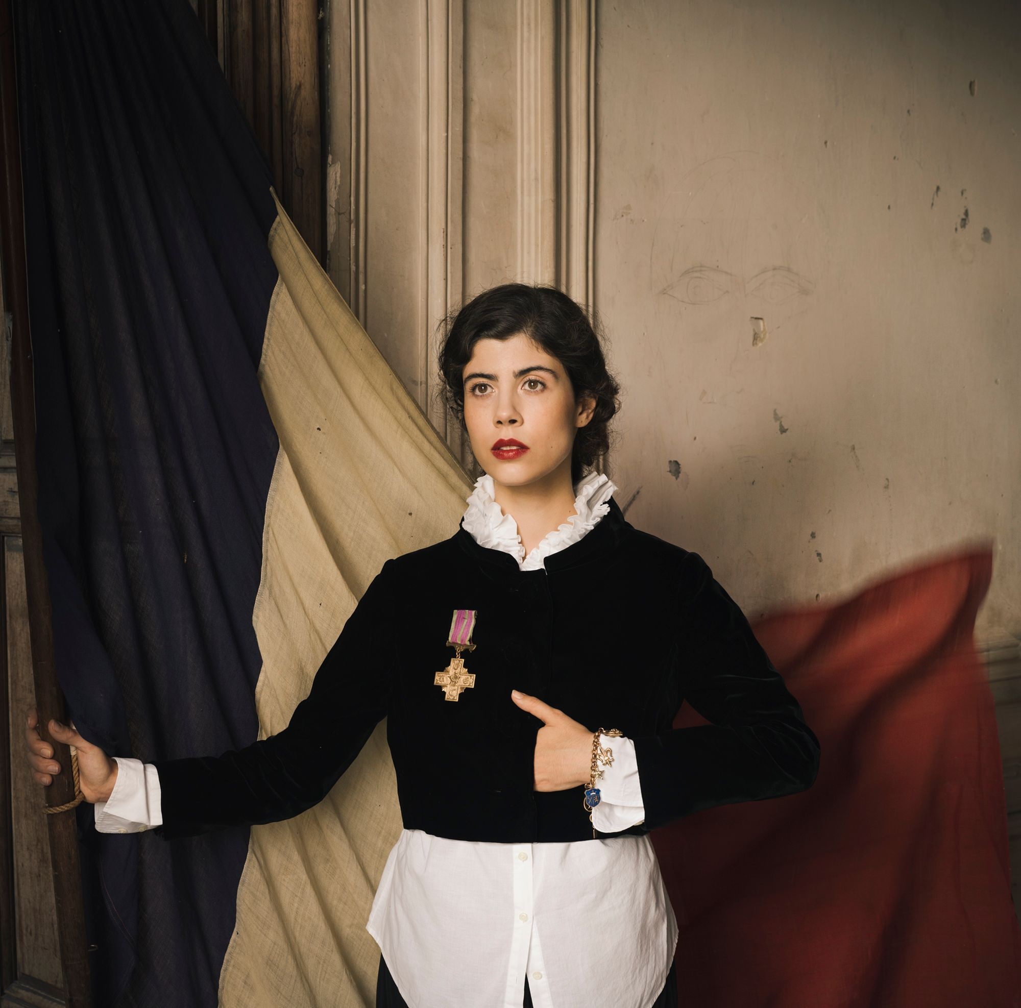 A dark-haired young woman stands facing the left, one hand tucked into a black velvet jacket with a French medal secured to the front. She holds a French flag behind her, a golden charm bracelet dangling from her other wrist. 