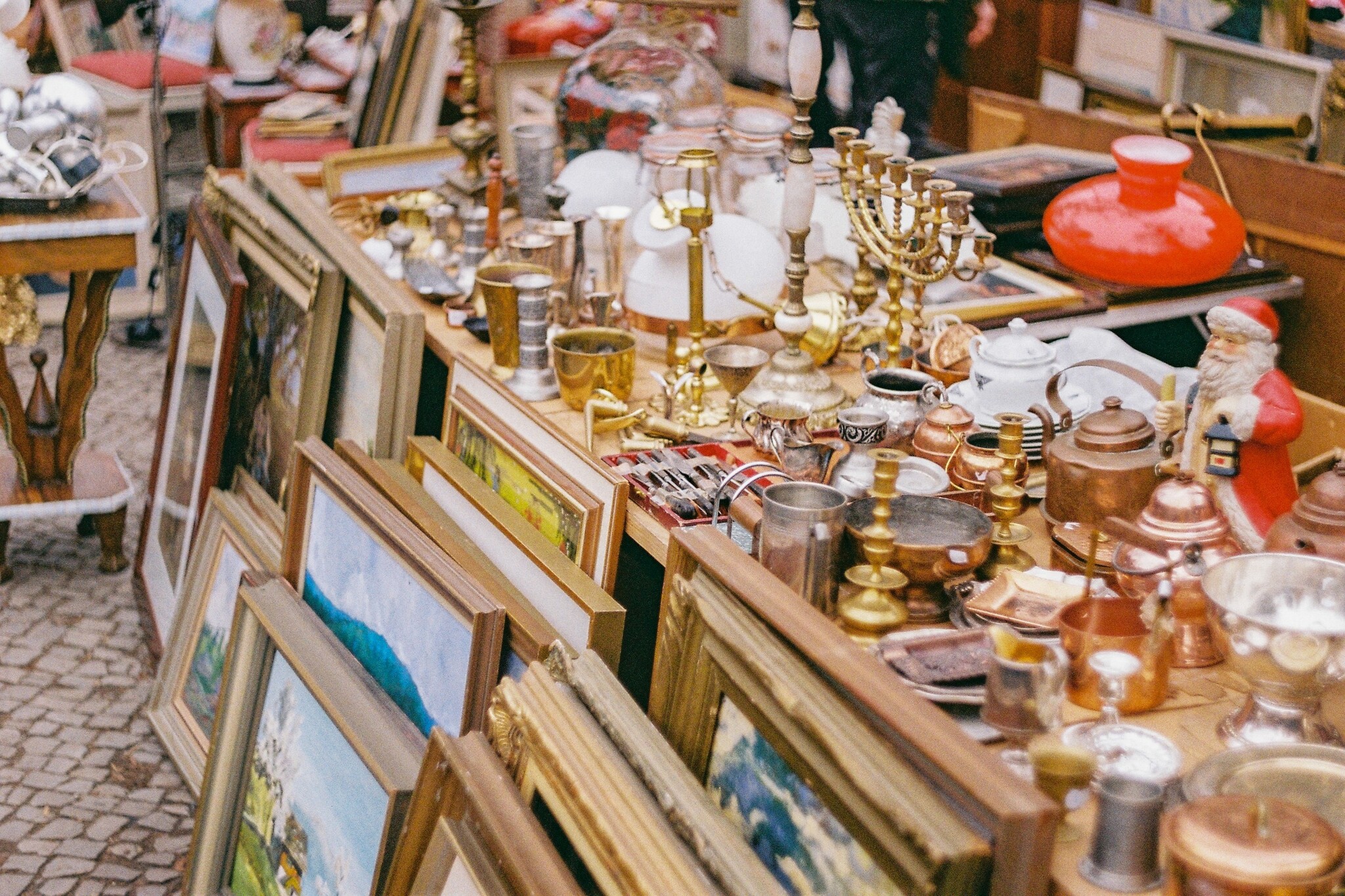 Stacks of paintings lean against a table laden with dishware, candlesticks, silver, and gold at an outdoor French flea market. It's colorful, inviting, and warm. 