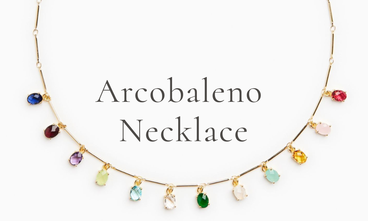 Our bright, colorful necklace on a light gold stick-chain hangs in a half-moon arc with 12 bright, glass stones representing each birth month. The words "Arcobaleno Necklace" hang above. 