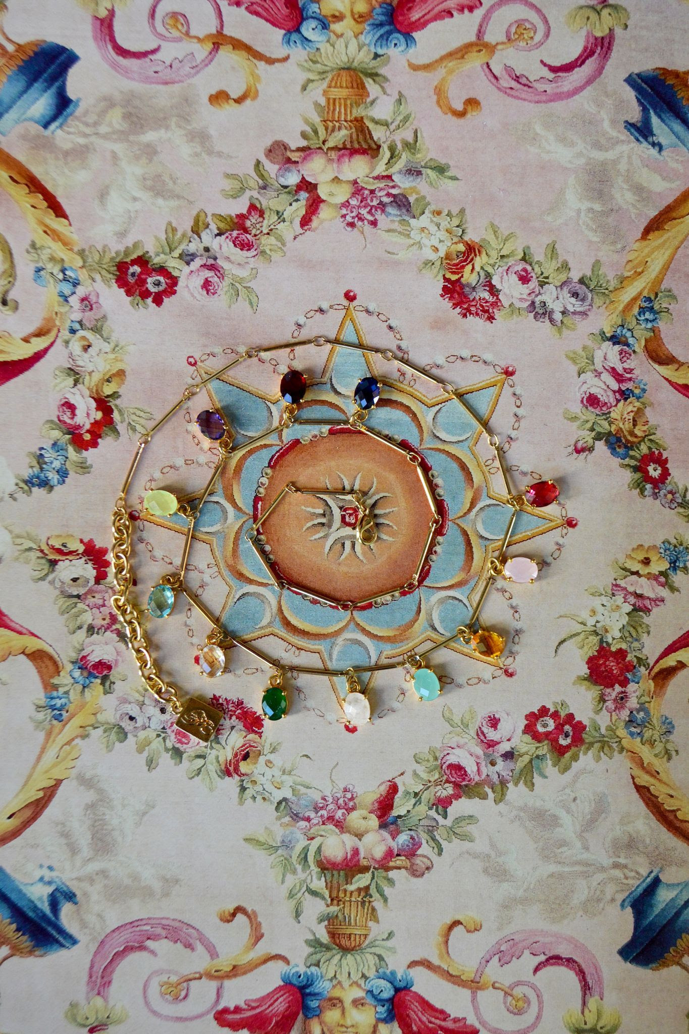 A colorful backdrop is painted like the European palace ceilings in blues, pinks, greens, and yellow. Spiraled around the center lies our golden stick-chain necklace which has 12 brilliant, glass stones attached at even intervals, the colors matching birthstones.