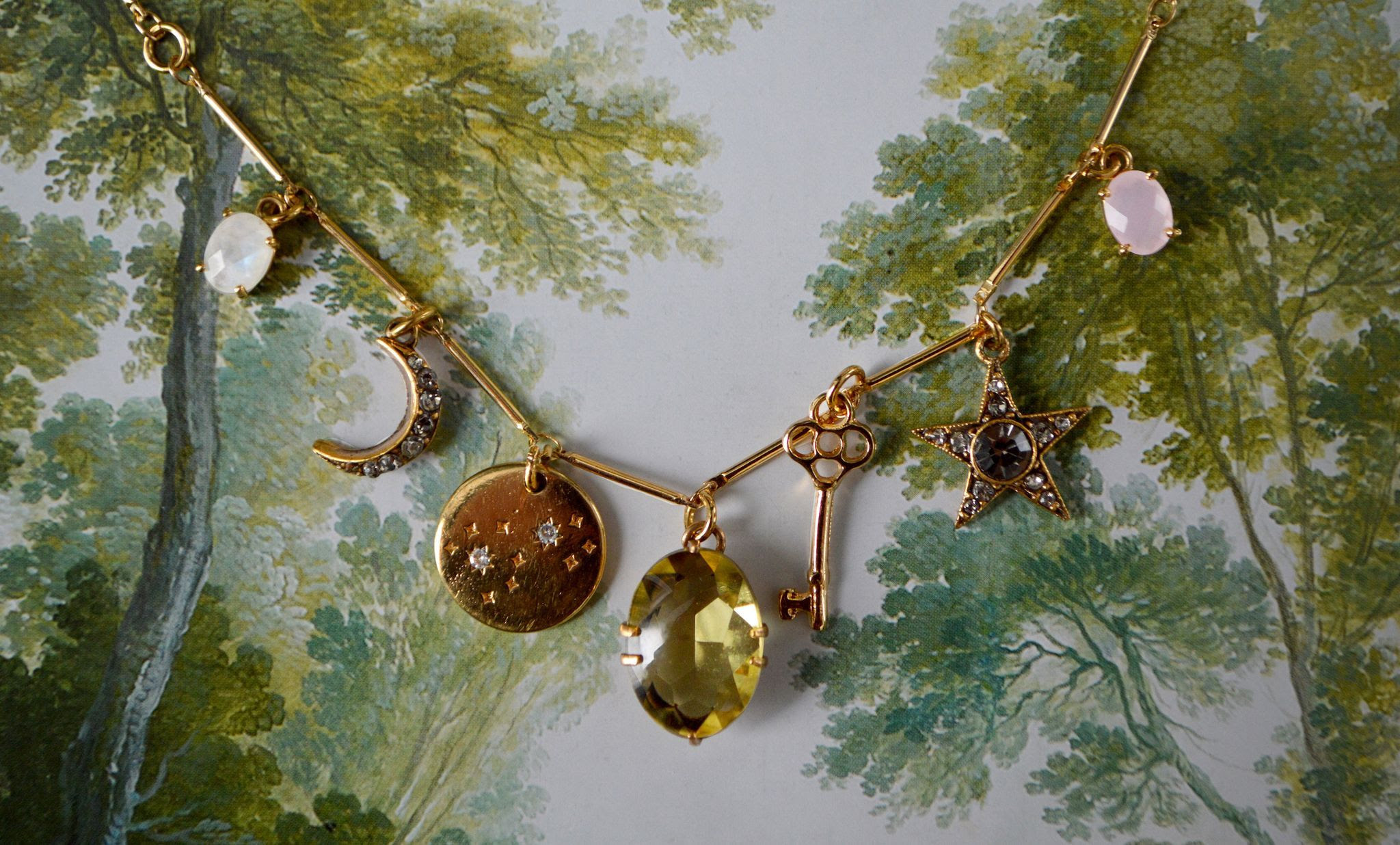 Laid on top of the painted backdrop of a gentle forest scene lies a light, golden stick chain necklace with charms spaced at even intervals—opalesque white, crescent moon, a gold circle with 2 petite stars, a sunshine-yellow central stone, golden key, star, and light pink stone charm.
