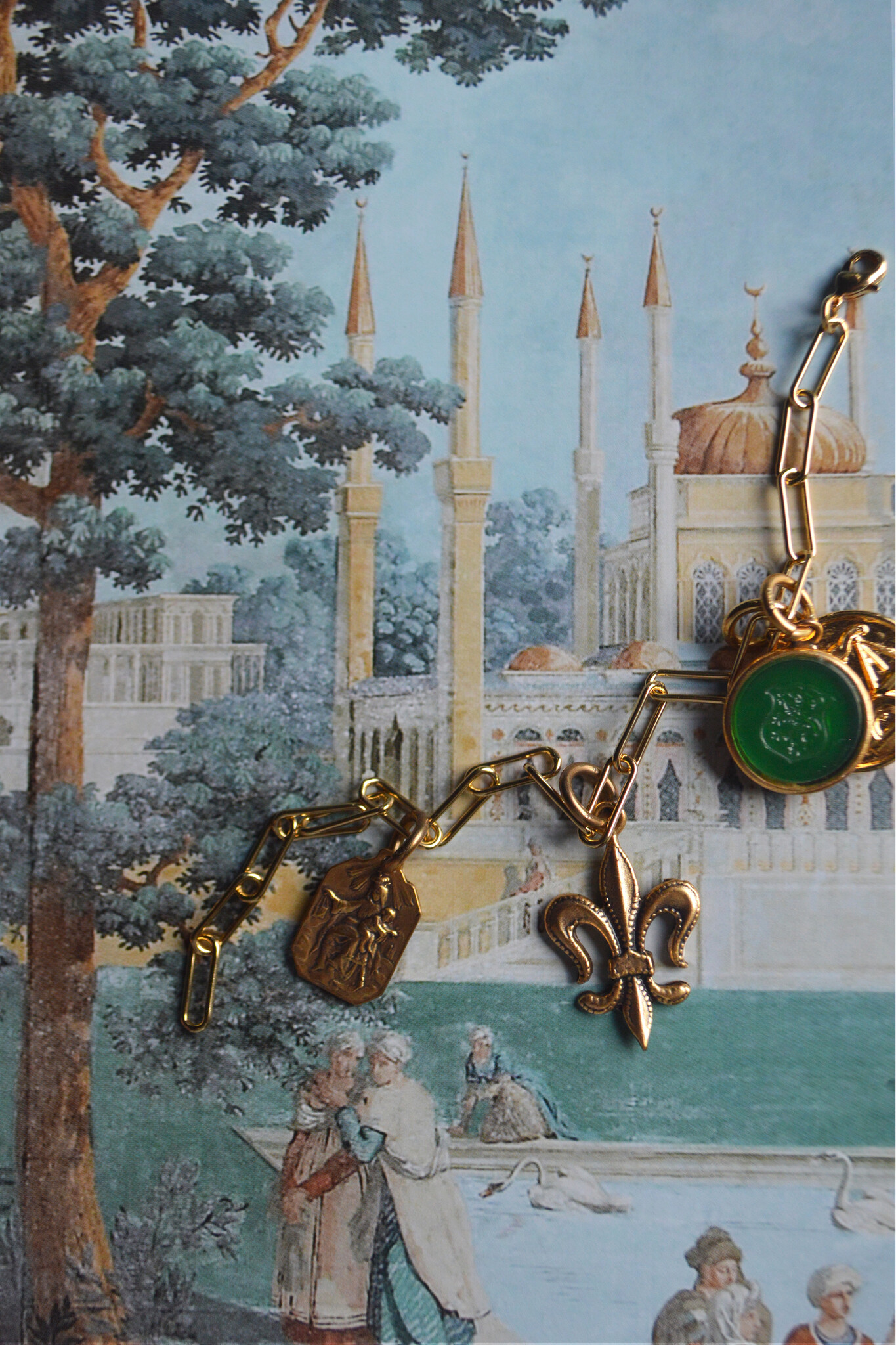 A paperclip chain drapes across a painted backdrop of a French court from the 18th century. A fleur-de-lis charm hangs from one part while a green, round intaglio charm drops from another, a golden Libra charm featuring raised scales, and a petite rectangular charm showcasing the holy virgin Mary with an infant Christ curled on her lap.