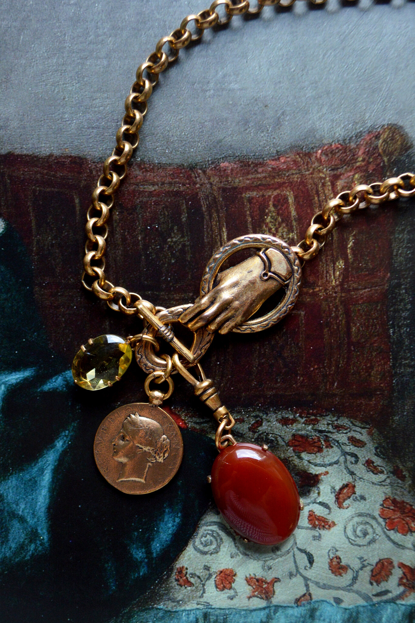 A golden hand holds onto a circle, from which dangle a deep red-orange oval stone (carnelian), a round golden coin with a woman's profile at center, and an oval transparent-yellow glass stone, all over a painted background in jewel tones.