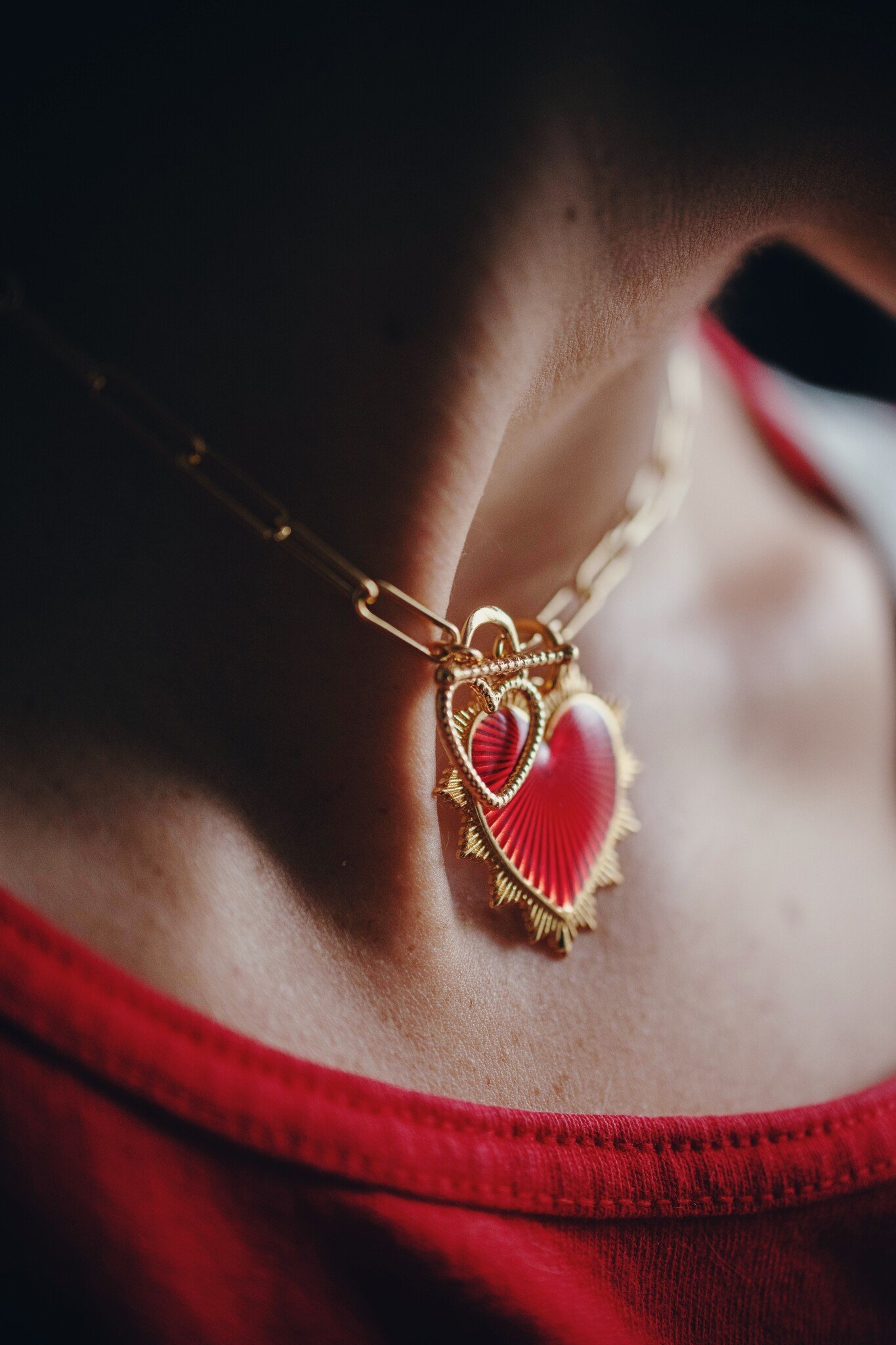 A bright red heart charm hangs from a golden, paperclip chain, with little beams radiating from its center to end in a series of points along the outer edge. 