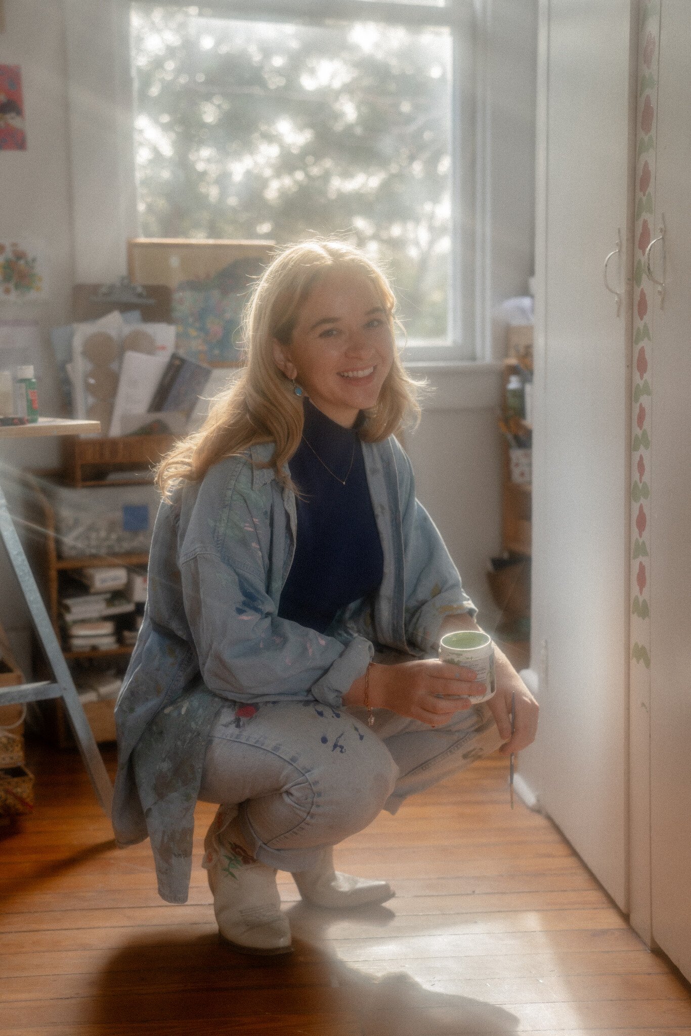 A young, white woman wearing light denim pants and jacket holds a coffee cup, smiling at the camera in hazy light.