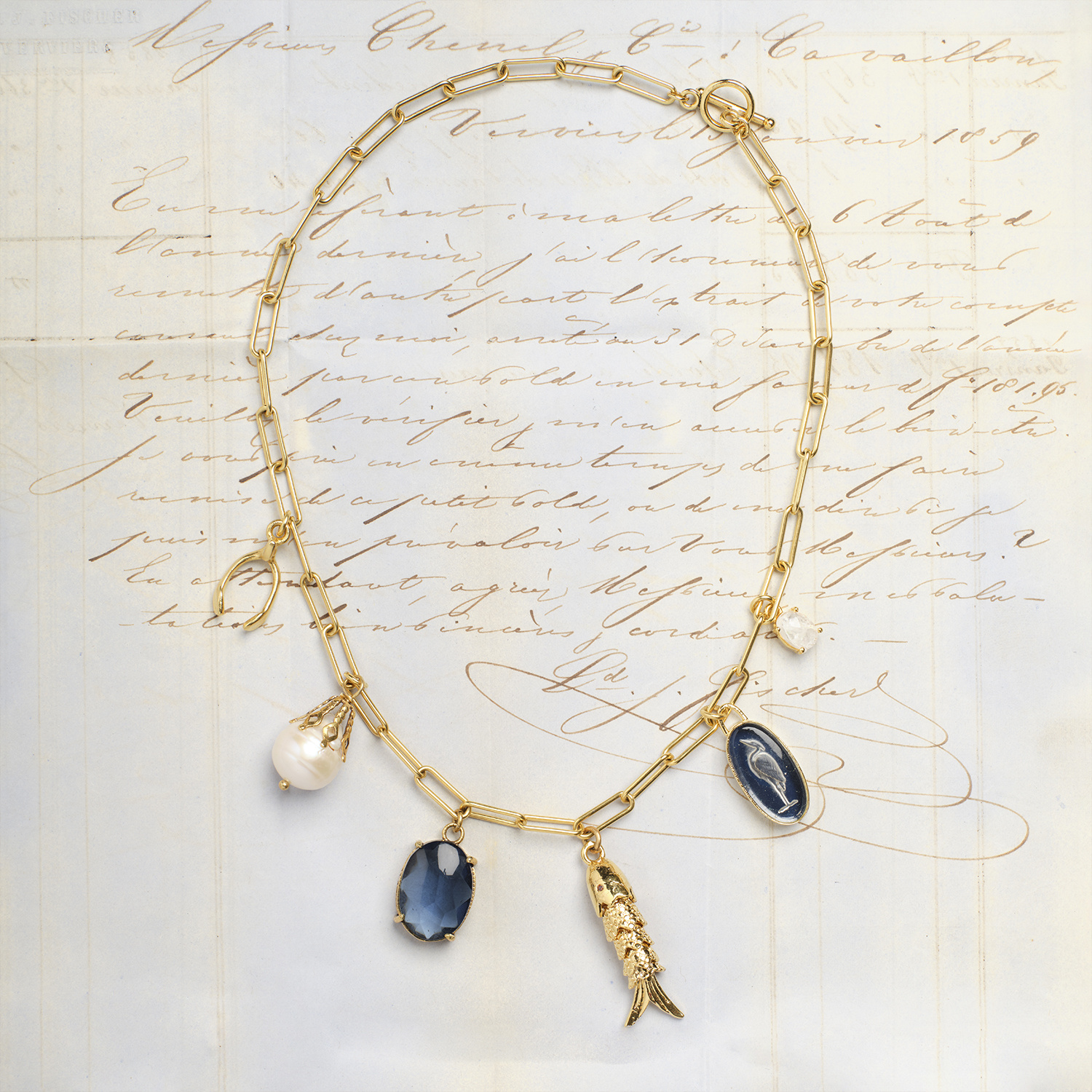 Over an antique French letter with script in sepia brown tones, a golden paperclip chain lies covered in blue, white, and gold charms around an aquatic theme.