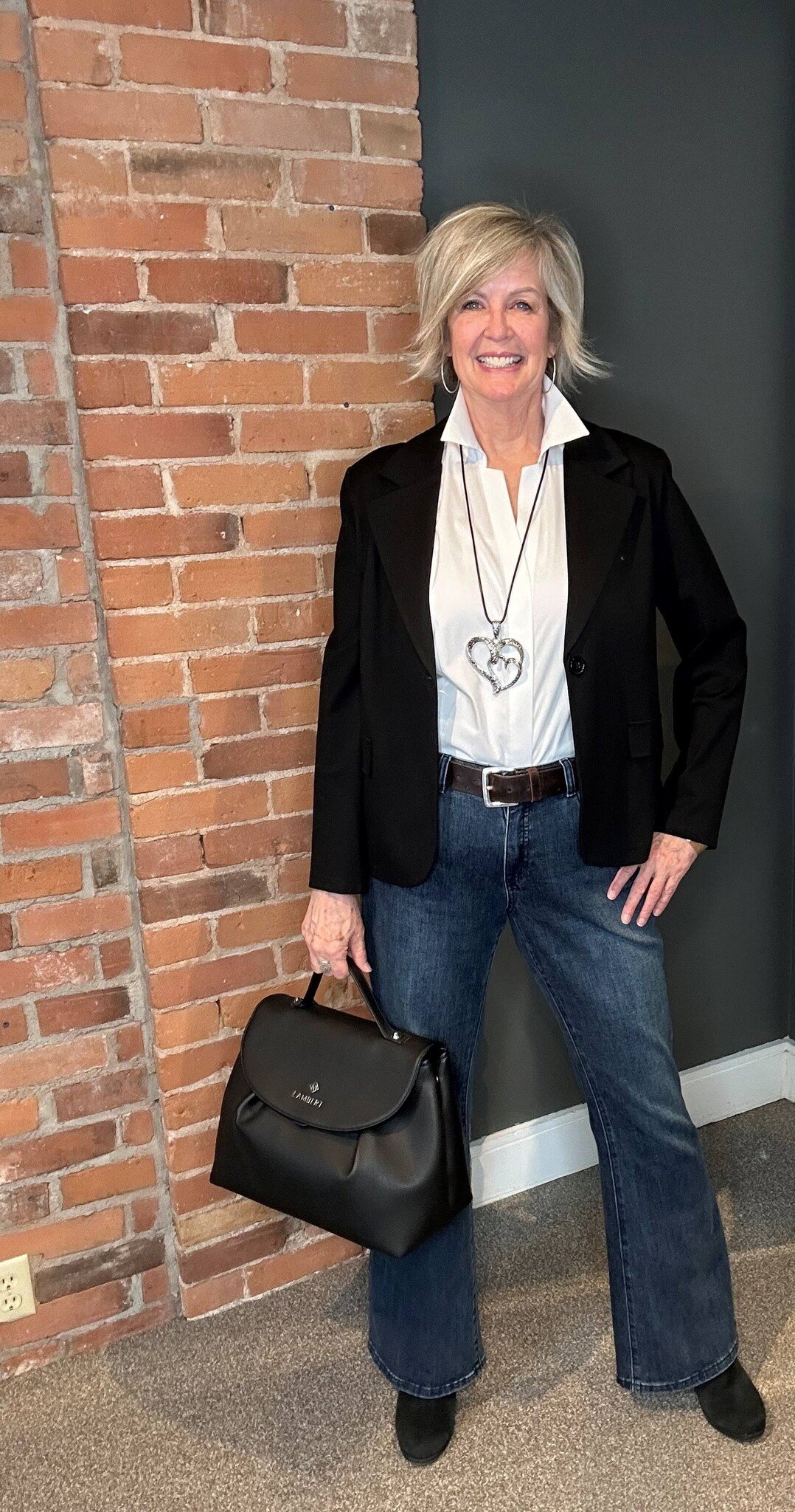 A woman wearing a pair of flared jeans, a white blouse, black blazer and holding a black purse.