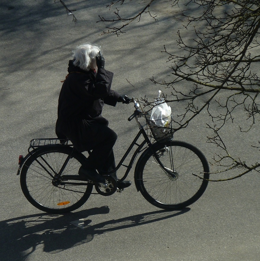 From slightly above we see a grey-haired woman riding a black step through bike on pavement.