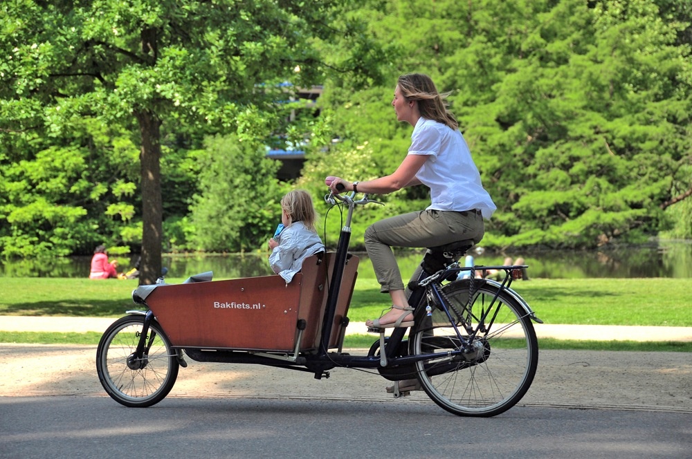 The sun shines on a casually dressed woman riding a two-wheeled box bike with a child in the front. They are riding on a park pathway next to a river. 