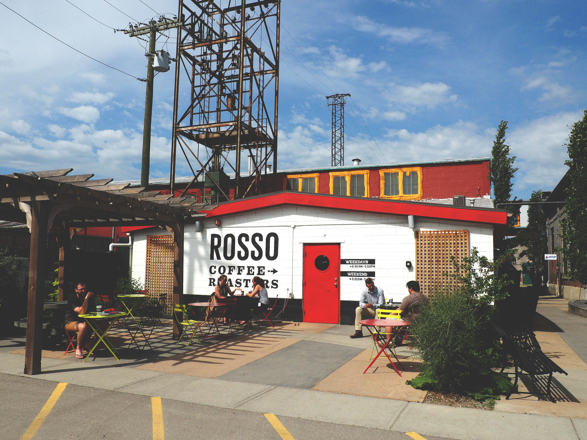 An outdoor shot of a red and white building with the Rosso logo on it. Guests enjoy coffee on folding tables and chairs on a semi covered patio out front.