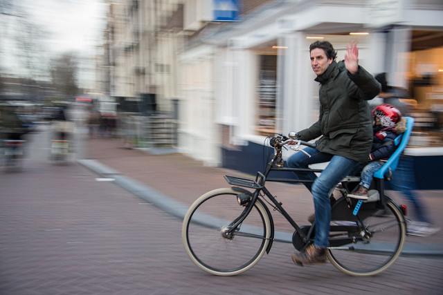 A man on a bicycle with a child on the rear child seat waves towards you.