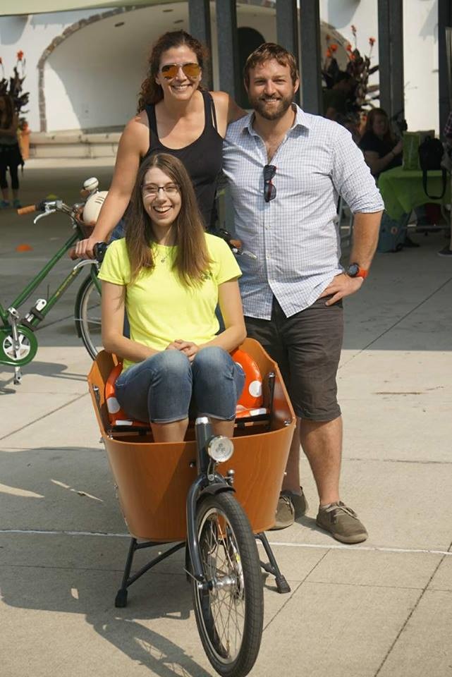 Ben and Caroline stand behind the Babboe bakfiets (box bike) in which Joy is sitting; they are all smiling.