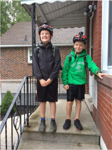 Two kids wearing bike helmets grin at the camera from their porch