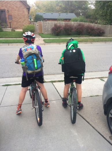 Two kids on bikes face the road from their driveway, ready for their commute to school.