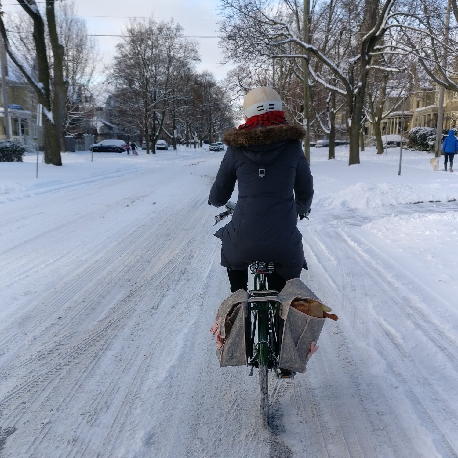 A woman rides away from the camera on a bike with panniers; the street is covered in packed snow.