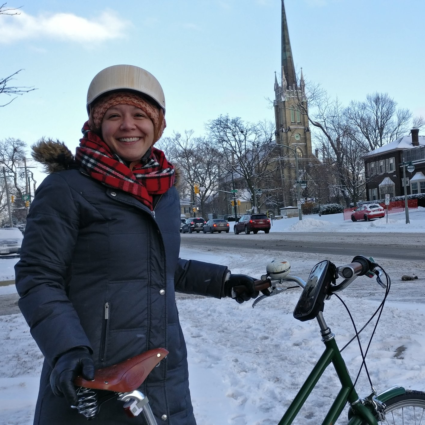 Caroline smiles at the camera, dressed for winter and holding her bike; a church spire and busy street are behind her.