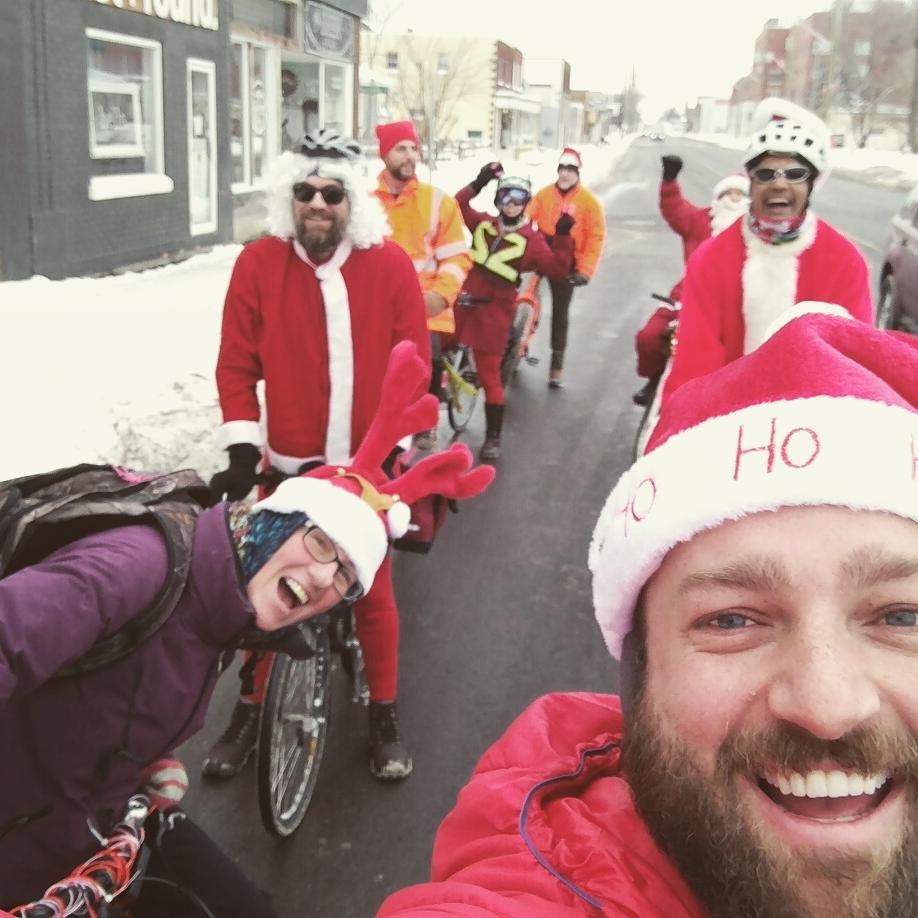 Ben wears a red coat and santa hat and takes a selfie over his shoulder at several similarly dressed people on their bikes.