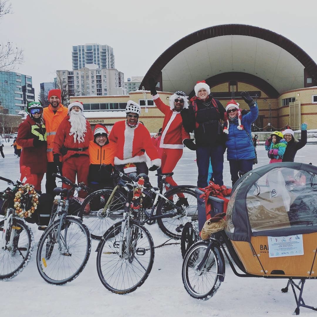 Cyclists dressed a bit like santas smile at the camera in front of an ice rink beyond which is a large outdoor bandshell.